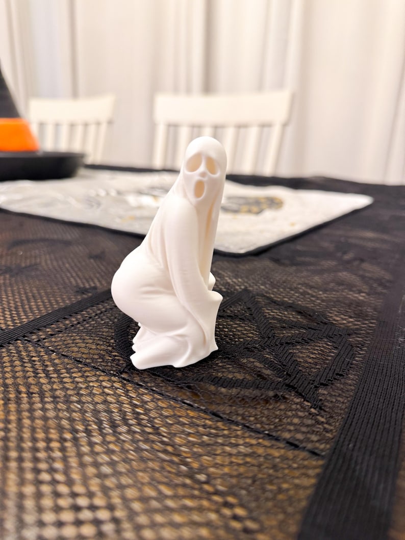 May include: A white ghost figurine in a kneeling pose, with a wide open mouth and eye sockets. The ghost is on a black mesh surface, with a table setting in the background. The figurine is a Halloween decoration.