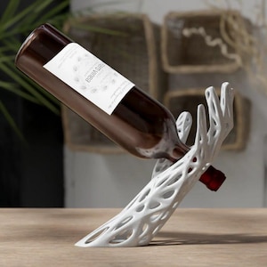May include: A white, abstract wine bottle holder in the shape of a hand cradles a brown wine bottle. The bottle has a white label with text. The holder is on a wooden surface.