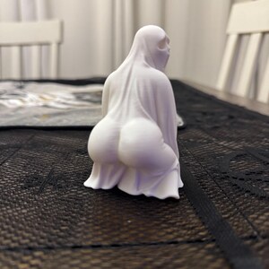 May include: A white ghost figurine in a squatting position. The ghost is draped in a sheet with a visible form underneath. The figurine has a simple, minimalist design, with a focus on the draped fabric and the implied shape beneath.