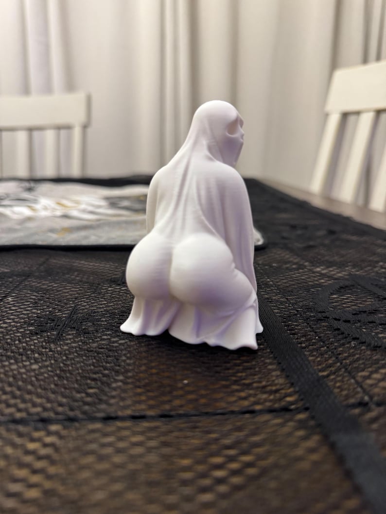 May include: A white ghost figurine in a squatting position. The ghost is draped in a sheet with a visible form underneath. The figurine has a simple, minimalist design, with a focus on the draped fabric and the implied shape beneath.