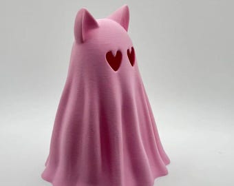 May include: A pink ghost-shaped figurine with cat ears and heart-shaped eye cutouts. The figurine has a draped design, giving it a flowing appearance. The color is a soft, pastel pink, and the overall design is whimsical.