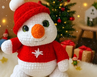 May include: A crocheted snowman figure, dressed in a red hat and sweater, with a white snowflake. It has an orange nose and green eyes. A small blue gift box with a red bow is in front. The background features a Christmas tree and festive decorations.