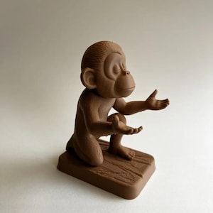 May include: A brown monkey figurine in a kneeling pose with its hands open. The monkey is on a rectangular base with a wood-grain texture. The figurine has closed eyes and a serene expression.