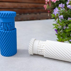 May include: Two cylindrical, decorative objects. One is bright blue, standing upright, and the other is white and lying on its side. Both have a textured, spiral design on the body and a geometric pattern on the top section. The background includes flowers.