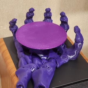 Könnte beinhalten: Ein lila, 3D-gedruckter, handförmiger Präsentationsständer mit einer kreisförmigen Plattform. Die Hand befindet sich in einer Greifposition, wobei die Plattform auf der offenen Handfläche und den Fingern ruht. Der Ständer ist für kleine Objekte konzipiert.