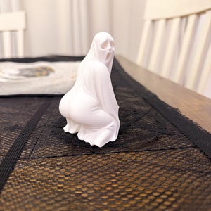 Può includere: Una statuetta di fantasma bianco in posizione accovacciata con la bocca aperta. Il fantasma è drappeggiato in un lenzuolo fluente, con i dettagli del viso e del corpo visibili. La statuetta è su un tavolo con un runner in pizzo nero.