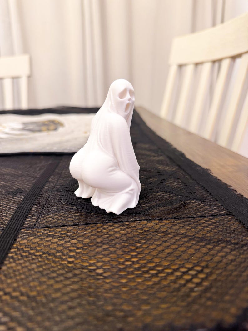 May include: A white ghost figurine in a squatting position with an open mouth. The ghost is draped in a flowing sheet, with the details of the face and body visible. The figurine is on a table with a black lace runner.