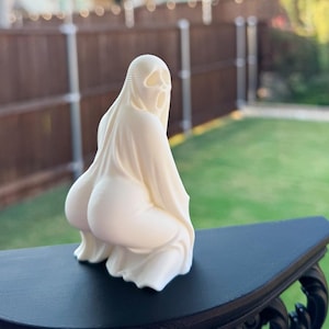 May include: A cream-colored sculpture of a figure in a draped garment, with a hood and facial features. The figure is kneeling, with the back and buttocks prominently displayed. The sculpture is on a black table.