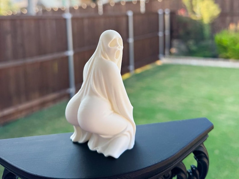May include: A cream-colored sculpture of a figure in a draped garment, with a hood and facial features. The figure is kneeling, with the back and buttocks prominently displayed. The sculpture is on a black table.