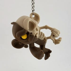 May include: A novelty keychain featuring a half-monkey, half-skeleton design. The monkey side is brown with a yellow eye, while the other half is a bone-colored skeleton. The keychain hangs from a silver chain.