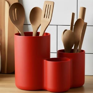 Op de afbeelding: Rode keukengerei-houder set met drie compartimenten, met houten lepels, spatels en ander keukengerei. De houders zijn cilindrisch en staan op een houten oppervlak, met een witte tegelachtergrond.