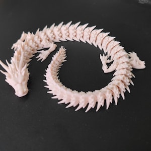 May include: A light pink, articulated dragon figurine. The dragon is made of many connected segments, allowing it to be posed. The head has detailed features, including horns and a defined snout. The dragon is lying on a dark surface.