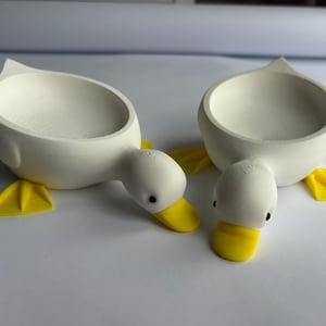 May include: Two white duck-shaped decorative bowls with yellow feet and beaks. The bowls have a rounded shape with a smooth, matte finish. The ducks are positioned on a white surface.