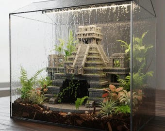 May include: A miniature terrarium featuring a detailed replica of a Mayan pyramid, surrounded by lush green plants and small colorful foliage. The glass enclosure has a black frame and is set on a wooden surface, creating a miniature ecosystem.