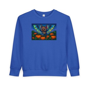 Puede incluir: Sudadera azul real con una colorida ilustración de un murciélago de dibujos animados con alas de arcoíris, sonriendo sobre un campo de calabazas naranjas. El diseño está centrado en la parte delantera de la parte superior de manga larga, ideal para Halloween.