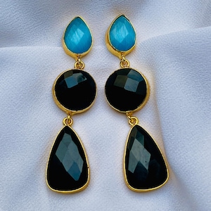 Pode incluir: Um par de brincos pendurados dourados. Cada brinco apresenta uma pedra preciosa azul em forma de gota na parte superior, uma pedra preciosa preta redonda no meio e uma pedra preciosa preta em forma de gota na parte inferior.
