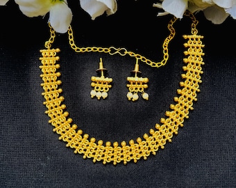 May include: A gold-colored necklace and earring set. The necklace and earrings have a detailed, textured design. The earrings have small, dangling white beads. The jewelry is displayed on a black surface, with white flowers in the background.