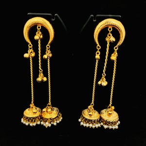 May include: A pair of gold-colored earrings with a crescent moon design at the top. The earrings feature dangling chains with small gold beads and bell-shaped pendants at the bottom, adorned with small white pearls. The background is black.