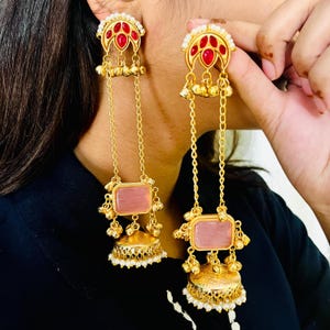 May include: Gold-toned earrings with red and pink accents. The earrings feature a floral design with dangling chains, pink rectangular stones, and small bell-shaped charms. The bottom of the earrings have a bell shape with white pearls.