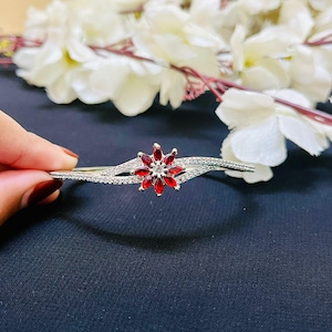 May include: A silver bracelet with a central flower design featuring red gemstones and a clear gemstone center. The bracelet has a delicate, curved design with small, clear gemstones along the band. The background includes white flowers.
