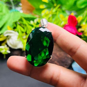 Puede incluir: Colgante de piedra preciosa verde esmeralda, de forma ovalada y facetada, con una fianza plateada. La piedra está engastada en un soporte plateado. El colgante se sostiene en una mano sobre un fondo borroso de follaje verde y una flor rosa.