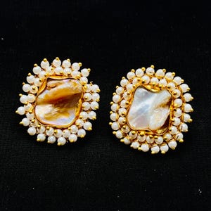 May include: A pair of gold-coloured stud earrings. Each earring has a large, irregularly shaped, iridescent pearl centre, surrounded by a halo of small, white, round pearls. The earrings are set against a black background.