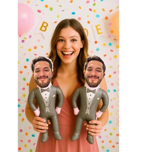 May include: A woman holding two inflatable dolls with a groom's face. The dolls are dressed in gray tuxedos with pink bow ties and cuffs. The background includes a banner that says "Bride" and colorful confetti.