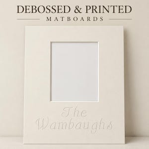 May include: A cream-colored rectangular matboard featuring a central rectangular cutout and the debossed text The Wambaughs below the opening. The top of the image displays the text DEBOSSED & PRINTED MATBOARDS in a dark brown serif font.