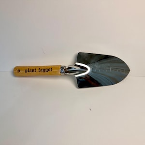 May include: A small, shiny metal garden trowel with a wooden handle. The handle has the text "plant faggot" printed on it. The trowel is designed for planting and gardening.