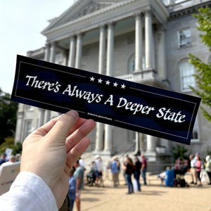 May include: A black bumper sticker with white text that reads "There's Always A Deeper State." The sticker has a dark blue background with a pattern and white stars. The sticker is held in front of a large building with columns and a crowd of people.