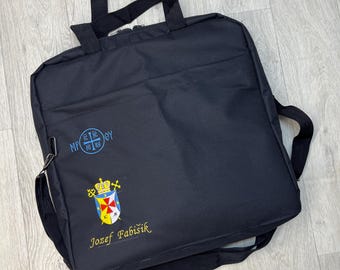 Puede incluir: Bolso tote negro con correa para el hombro y asas superiores. La bolsa presenta texto bordado y un escudo con una corona, una cruz y un escudo en amarillo, rojo, azul y blanco. El texto "Jozef Fabisik" está bordado en oro.