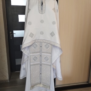 May include: White religious garment with intricate silver embroidery. The garment features a high collar, long sleeves, and a decorative panel down the front. The embroidery includes geometric and floral patterns. The garment is displayed on a black mannequin head.