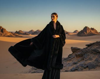 May include: A long, flowing black cloak featuring a thick, dark faux fur trim along the collar, front opening, and hemline. The garment is constructed from a smooth, solid black fabric that drapes to the floor, creating a wide, dramatic silhouette.