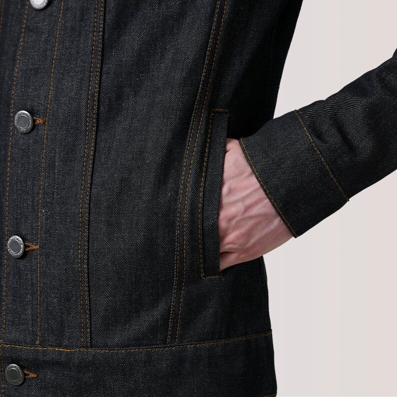May include: A dark denim jacket with visible orange stitching and silver buttons. The jacket has a pocket on the chest and the person's hand is in the pocket. The jacket is buttoned up.