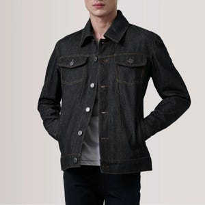 May include: A dark denim jacket with a classic collar, button-up front, and two chest pockets. The jacket features orange stitching and silver buttons. The model is wearing the jacket with a white t-shirt and dark blue pants.