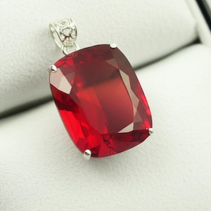 Op de afbeelding: Een rechthoekige, gefacetteerde rode edelsteen hanger met een zilveren beugel. De edelsteen is dieprood en vangt het licht. De hanger is gezet in een zilveren zetting met kleine pootjes. De beugel heeft een decoratief ontwerp.