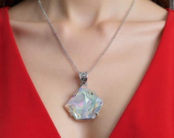 May include: A silver necklace featuring a large, iridescent, geometric pendant. The pendant has a multifaceted, crystal-like appearance, reflecting a spectrum of colors. The necklace is worn against a red background.