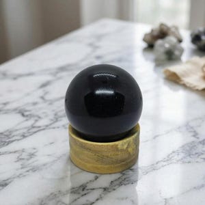 Könnte beinhalten: Eine polierte schwarze Obsidiankugel, etwa 7.6 cm im Durchmesser, auf einem Naturholzständer. Die Kugel reflektiert das Licht und zeigt ihre glatte, glänzende Oberfläche. Der Ständer hat eine hellbraune Farbe.