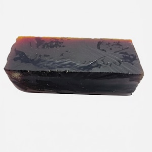 May include: A dark, rectangular stone with a textured surface. The top edge has a reddish-brown colour, contrasting with the darker body. The stone appears to be a natural, raw material, possibly a mineral or gemstone.
