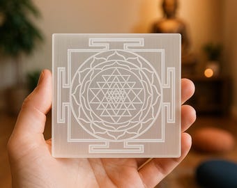 Puede incluir: Placa cuadrada de cristal de selenita blanco translúcido con un diseño Sri Yantra grabado. El patrón geométrico presenta triángulos entrelazados y un diseño central de flor de loto. La placa se sostiene en una mano, con un fondo borroso.