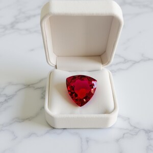 Peut inclure: Une grosse pierre précieuse rouge rubis, taillée en forme de triangle, repose dans un écrin blanc ouvert. La boîte est doublée d'un matériau blanc doux, sur un fond marbré.