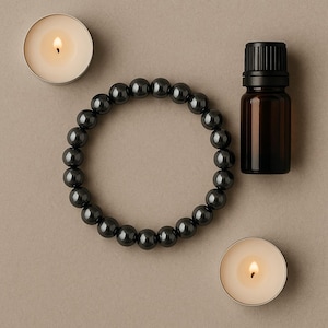 May include: A top-down view of a wellness arrangement featuring a dark gray beaded bracelet, a small amber glass bottle with a black cap, and two lit tea light candles. The items are arranged on a neutral-toned surface.