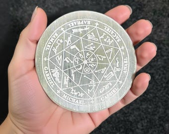 May include: A silver-colored, circular decorative item with etched symbols and text. The item is held in a hand and features a central star-like design surrounded by names and other markings. The overall aesthetic is mystical.
