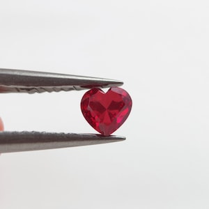 May include: A faceted, heart-shaped, deep red gemstone is held by a pair of tweezers. The gem has multiple facets that reflect light, creating a sparkling effect. The tweezers are silver-colored and hold the gem against a white background.