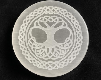 Peut inclure: Plaque ronde en cristal de sélénite blanche, gravée d'un motif d'arbre de vie. L'arbre est entouré d'une bordure de nœuds celtiques. La plaque a une surface lisse et est posée sur un fond sombre.