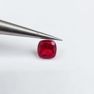 May include: A vibrant, cushion-cut ruby gemstone, held by a pair of tweezers. The ruby is a deep, saturated red color, with facets that catch the light. The tweezers are silver-colored, and the background is plain white.