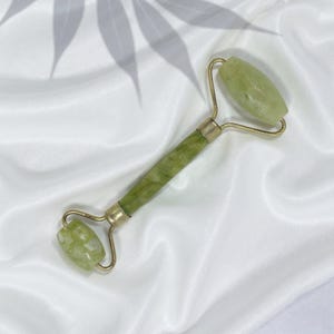 Op de afbeelding: Een jade gezichtsroller met een goudkleurig metalen frame. De roller heeft aan elk uiteinde twee afgeronde jadestenen en een groen en wit gemarmerd handvat. De achtergrond is een witte, zijdeachtige stof met een schaduw van een blad.
