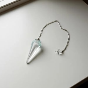 Puede incluir: Un péndulo de cristal transparente con punta y cadena plateada. El cristal tiene un tono azul claro en la parte superior. La cadena termina con una pequeña cuenta de cristal transparente. Diseñado para la adivinación y prácticas espirituales.
