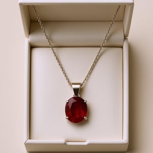May include: A silver necklace with a large, oval, deep red gemstone pendant. The pendant is set in a silver bezel and is displayed in an open white jewelry box with a cream-colored interior.