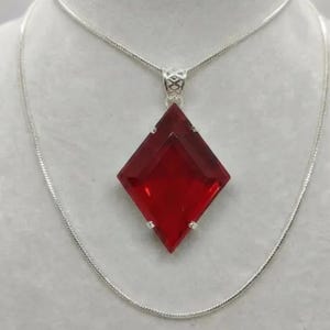 Op de afbeelding: Een zilveren ketting met een grote, rode, ruitvormige hanger. De hanger is een gefacetteerde edelsteen met een helder, reflecterend oppervlak. De ketting is een fijne, zilveren slangenketting.